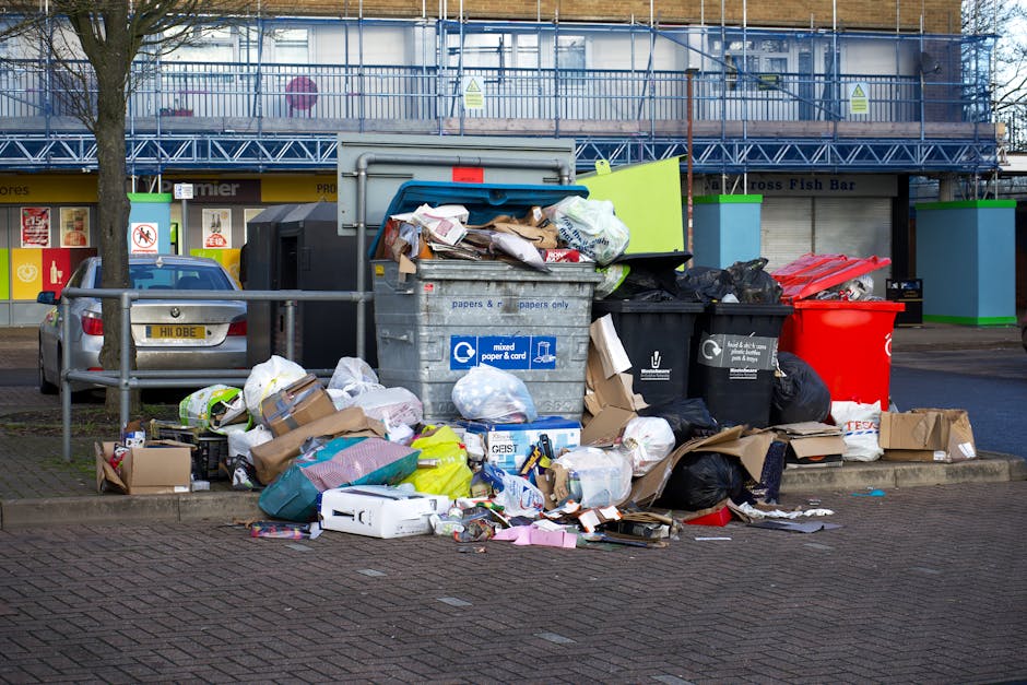 A pile of black bin bags and discarded packaging materials, including cardboard and foam, are stacked haphazardly in front of a red metal door embedded in a beige textured concrete wall. The trash app